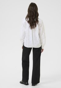 Chemise blanche à boutons avec des accents à motifs, associée à un pantalon noir ample et des chaussures noires plates. Tissu lisse, manches longues.