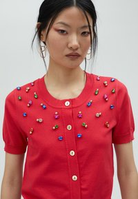 Cardigan en tricot rouge avec des manches courtes, un col rond, et des gems et perles décoratives sur le devant. Fermeture à boutons avec des boutons assortis.
