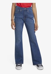 Jeans en denim bleu évasés avec une taille haute, un design à cinq poches et des détails de couture contrastants. Portés avec des baskets noires et blanches.