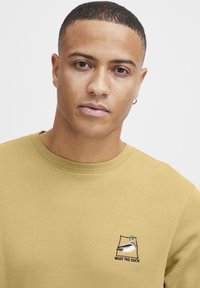 Mustard-yellow sweatshirt with round neckline, featuring an embroidered duck graphic and text "WHAT THE DUCK" on the chest.
