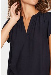 Donna con capelli ricci che indossa una blusa corta a maniche corte di colore scuro e con scollatura a V, mano alzata vicino all'orecchio.