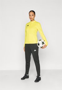 Κίτρινο μπλουζάκι με φερμουάρ και μαύρες λεπτομέρειες, σε συνδυασμό με μαύρο αθλητικό παντελόνι και μαύρα αθλητικά παπούτσια. Κρατάει μια ασπρόμαυρη μπάλα ποδοσφαίρου.