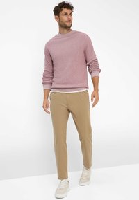 Lichtroze getextureerde trui met geribbelde zoom, gecombineerd met beige taperende broek en witte sneakers. Model staat voor een neutrale achtergrond.