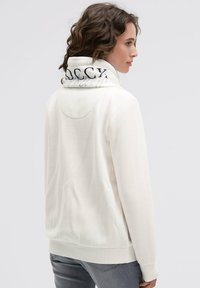 Cream-colored sweatshirt with a high collar featuring a black logo print. Soft texture, relaxed fit, and minimalistic design.