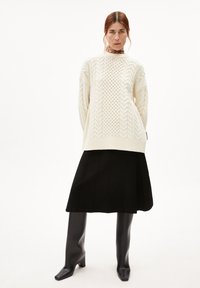 Crème-kleurige kabelgebreide trui met een losse pasvorm, gecombineerd met een zwarte plissérok en zwart hoge laarzen met hak.