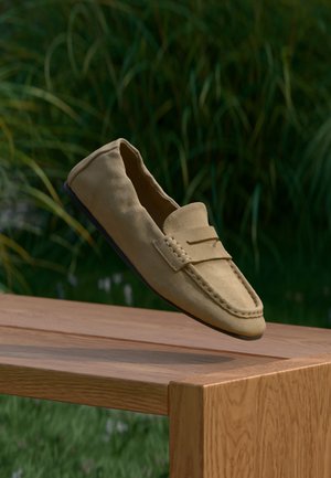 Ταμπά σουέντ μοκασίνι με στρογγυλεμένη μύτη, διακοσμητικό ράψιμο και μαύρη επίπεδη σόλα, κρεμασμένο πάνω από μια ξύλινη επιφάνεια με πράσινο φόντο.