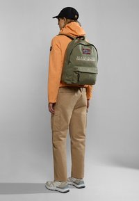 Zelený batoh s logem a detailem vlajky, oranžová bunda, béžové cargo kalhoty a tenisky. Batoh má přední kapsu a stahovací uzávěr.
