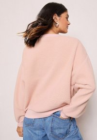 Maglione in pile rosa chiaro con una finitura morbida e texture. Presenta polsini e orlo a costine e una vestibilità rilassata e oversize.