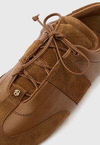 Καφέ δερμάτινο και σουέτ αθλητικό παπούτσι με δεμένα κορδόνια και μικρή χρυσή λεπτομέρεια λογότυπου στο εμπρόσθιο μέρος, απεικονιζόμενο σε λευκό φόντο.