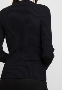 Pull noir côtelé à manches longues, avec un col arrondi et un tissu texturé. Comprend des coutures discrètes le long du dos.