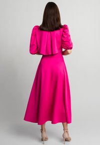 Kobieta stojąca tyłem, ubrana w jasnoróżową sukienkę z bufkami oraz beżowe sandały na obcasie, na jednolitym szarym tle.