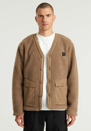 Brun fleece cardigan, strikket tekstur, knaplukning, ribbede kanter, to forlommer, kontrasterende stofdetaljer, logo-patch på brystet.