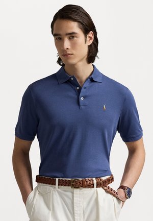 Navyblauwe polo met een kraag, korte mouwen en een klein geborduurd logo. Bruine gevlochten riem, beige broek en een zichtbare horloge.