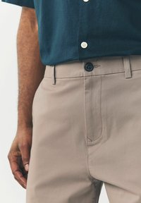 Beige nadrágot viselő férfi, fekete gombbal és sötétkék, rövid ujjú inggel, amely be van tűrve, áll, egyik keze lazán a teste mellett.