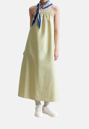Persoon draagt een mouwloze lichtgroene maxidress, witte sneakers, beige sokken en een blauw-wit patroon neksjaal tegen een witte achtergrond.