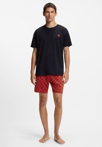 Melns kokvilnas T-krekls ar mazu sarkanu logotipu, kombinēts ar sarkaniem peldēšanas šortiem, kuros ir atkārtots grafiskais raksts melnā un oranžā krāsā.