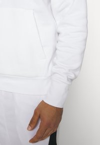 Felpa bianca con tasca frontale, polsini a coste e finitura testurizzata, abbinata a pantaloni leggeri dello stesso colore. Mano visibile sul lato.