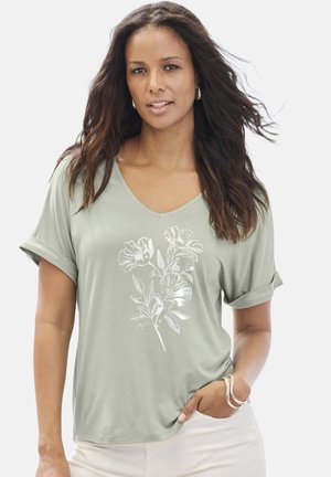 Frau mit langem dunklem Haar trägt ein hellgrünes V-Ausschnitt-Shirt mit weißem Blumenmuster, weiße Hose sowie silberne Creolen und Armbänder.