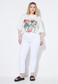 Vita denimjeans i kombination med en oversized ljusbeige t-shirt med en färgglad Rolling Stones-grafik och svarta slip-on sandaler.