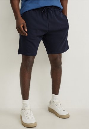 Mand iført marineblå shorts med snøre, hvide sokker og hvide sneakers står mod en ensfarvet baggrund.