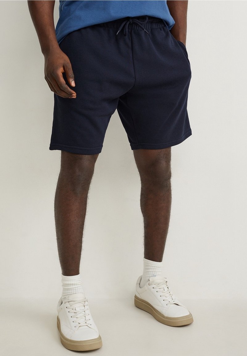Homme portant un short bleu marine à cordon, des chaussettes blanches et des baskets blanches, debout devant un fond uni.