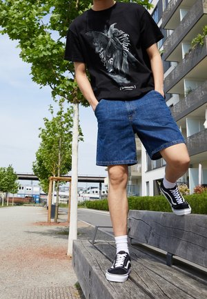 Person in schwarzem Grafik-T-Shirt, blauen Jeansshorts und schwarzen Sneakers steht mit einem Fuß auf einer Holzbank in einem urbanen Außenbereich.