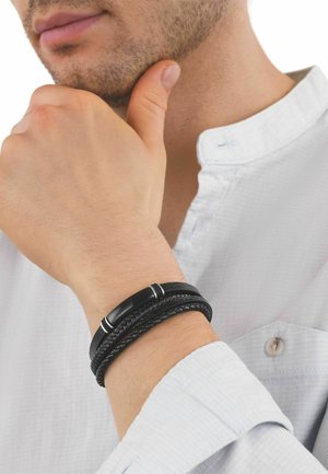 Uomo con camicia bianca e bracciale di pelle intrecciata nero, con la mano appoggiata sul mento, che mostra barba e labbra in primo piano.