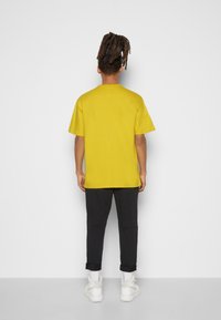 Žluté bavlněné tričko s krátkými rukávy, volný střih a kulatým výstřihem, spárované s černými kalhotami a bílými teniskami.