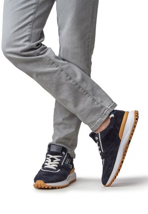 Marineblauwe suède sneakers met witte veters en een rubberen zool met een beige accent. Gecombineerd met lichtgrijze slim-fit jeans.