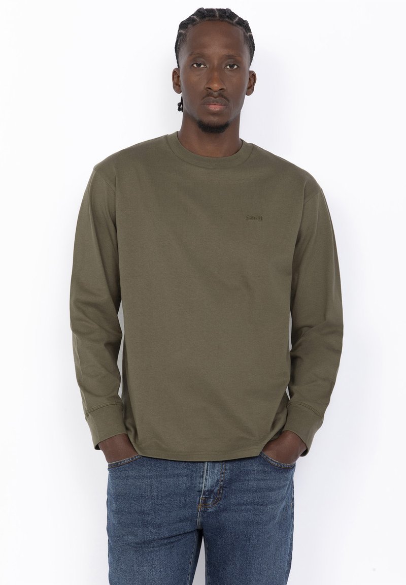 Sweatshirt vert olive à manches longues en coton, avec un col rond et des poignets cousus, associé à un jean en denim bleu.