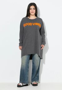 Túnica gris de manga larga con "GOOD VIBES" en naranja brillante. Combinada con jeans de pierna ancha de un azul deslavado y sandalias de plataforma con acentos plateados.