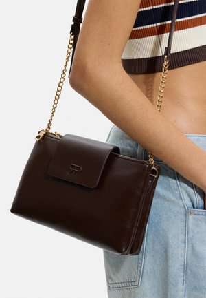 Ciemnobrązowa skórzana torba na ramię ze złotym łańcuszkiem, noszona przez osobę w paskowanym krótkim topie i jasnoniebieskich dżinsach.