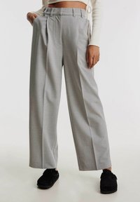 Grijze, hoge taille, geplooide broek met brede pijpen en riemlussen, gemaakt van een gestructureerde stof. Gecombineerd met zwarte schoenen.