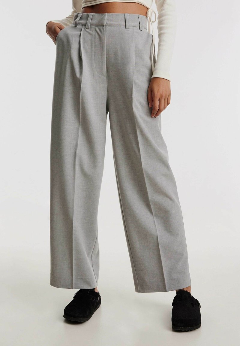 Grijze, hoge taille, geplooide broek met brede pijpen en riemlussen, gemaakt van een gestructureerde stof. Gecombineerd met zwarte schoenen.