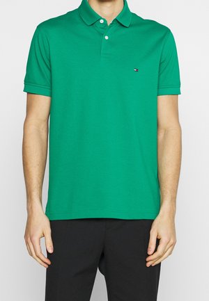 Mand iført en grøn kortærmet polo med et lille logo på brystet og sorte bukser, stående mod en ensfarvet baggrund.