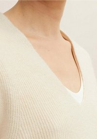 V-ringad tröja i mjuk beige stickning, med ett texturerat mönster och en avslappnad passform, över en vit topp. Närbild av halslinningen.