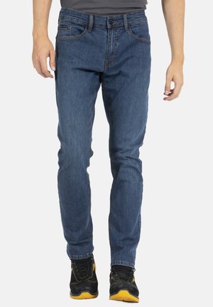 Jean en denim bleu avec une coupe droite, comportant cinq poches, des détails cousus et une fermeture à bouton. Porté avec des chaussures noires et jaunes.