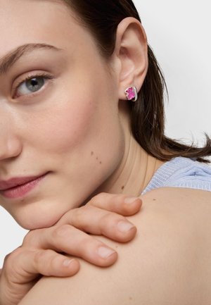 Joven mujer apoyando la barbilla sobre las manos cruzadas, lleva un pendiente plateado con diseño de flor rosa, se ve una blusa azul claro en el hombro.