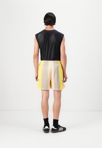 Svart ermeløs topp kombinert med beige og gule nettingshorts. Shortsene har en avslappet passform og elastisk midje. Sorte sokker og sko.