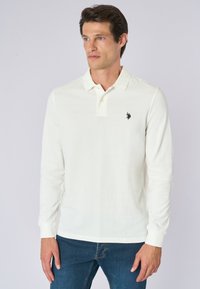 Polo a maniche lunghe bianco realizzato in tessuto morbido, con colletto classico, patta con tre bottoni e un piccolo logo nero sul petto.
