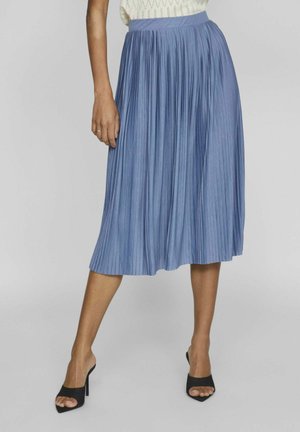 Woman wearing a blue pleated midi skirt, white sleeveless top, and black open-toe high heels, standing with one leg slightly forward.