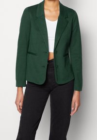 Blazer vert en tissu texturé, avec un col cranté, deux boutons et une coupe ajustée. Porté sur un haut blanc et associé à un pantalon noir.