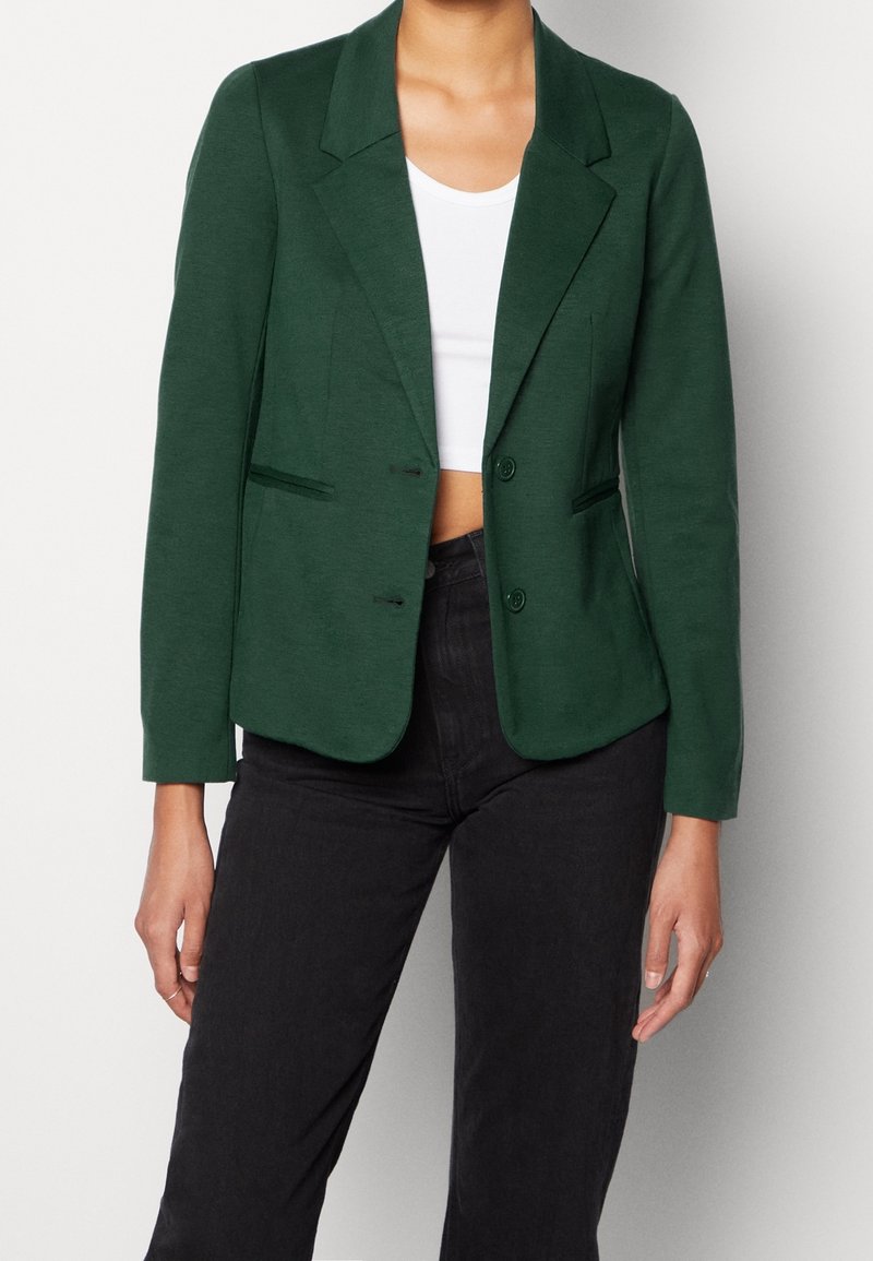 Blazer vert en tissu texturé, avec un col cranté, deux boutons et une coupe ajustée. Porté sur un haut blanc et associé à un pantalon noir.