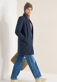 Manteau long matelassé bleu marine avec poches latérales, associé à un col roulé marron, un jean large bleu et des baskets beige, tenant un sac vert olive.