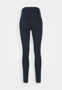 Jean skinny en denim foncé, avec une taille haute, un design cinq poches et une couture discrète sur les poches arrière. Texture du tissu lisse.