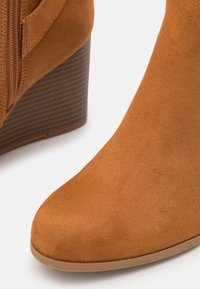 Stivale ankle in suede marrone con tacco a cuneo, punta arrotondata, zip laterale e tacco a righe. Texture liscia con dettagli minimi.
