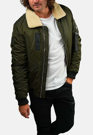 Homme portant une veste bomber verte avec col en peau retournée, t-shirt blanc, pantalon noir, posant avec une main dans la poche devant un fond uni.