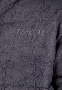 Grått, texturerat tyg med en skrynklig yta; har "KANI" broderat i en mörkare grå färg, subtil detaljer på ytan.