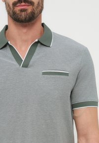 Άνδρας που φοράει γκρι πόλο πουκάμισο με πράσινη και λευκή διακόσμηση στο γιακά, V-λαιμόκοψη, τσέπη στο στήθος και μανσέτα στο μανίκι, εμφανίζει τον άνω κορμό και γένια.