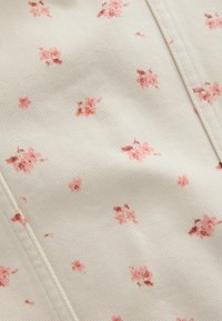 Tissu crème avec de petites impressions florales roses dispersées et des lignes de couture diagonales visibles.